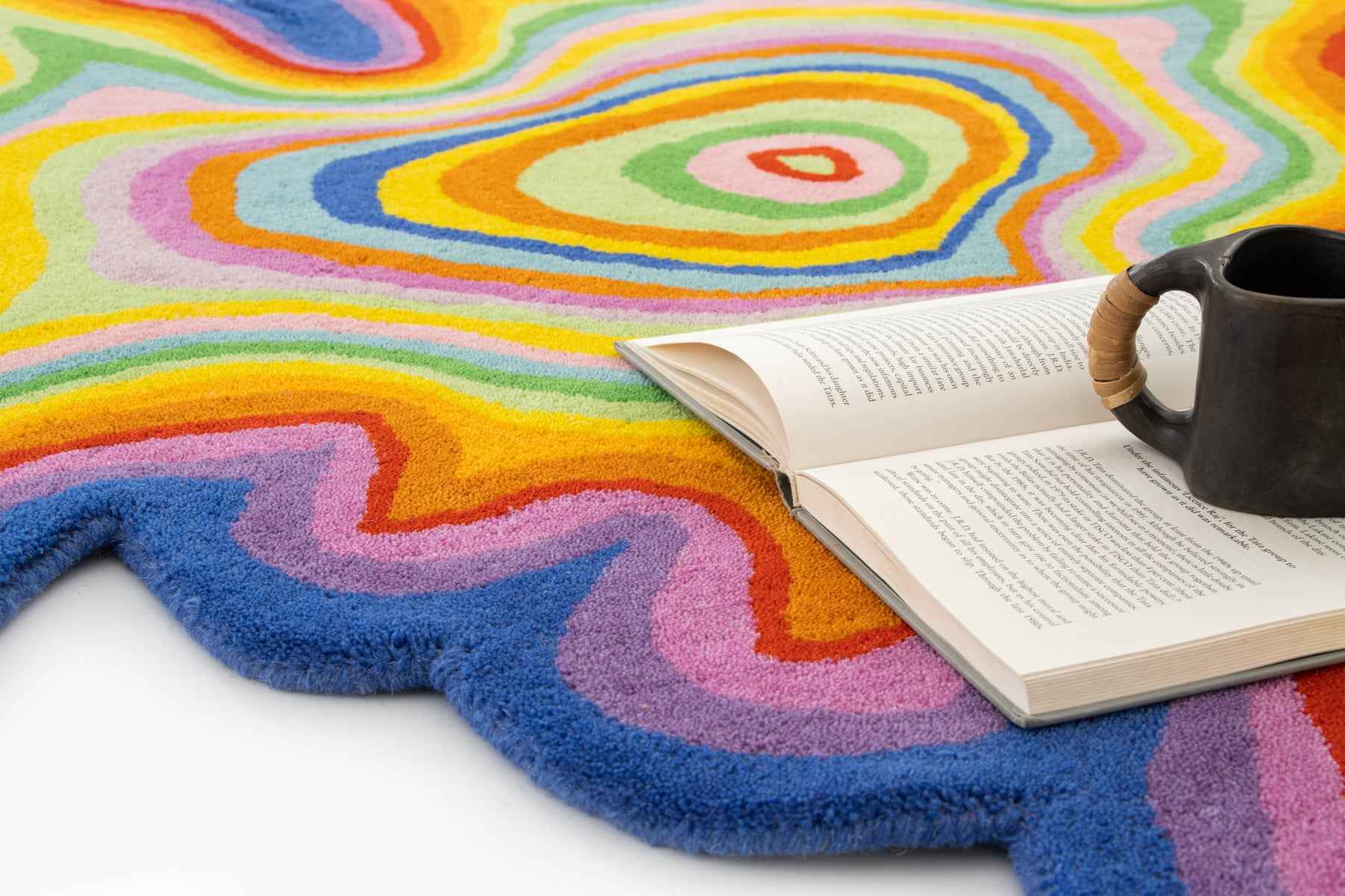 Psychedelic rainbow wool rug with open book and ceramic mug on white surface | Beyond Rugs