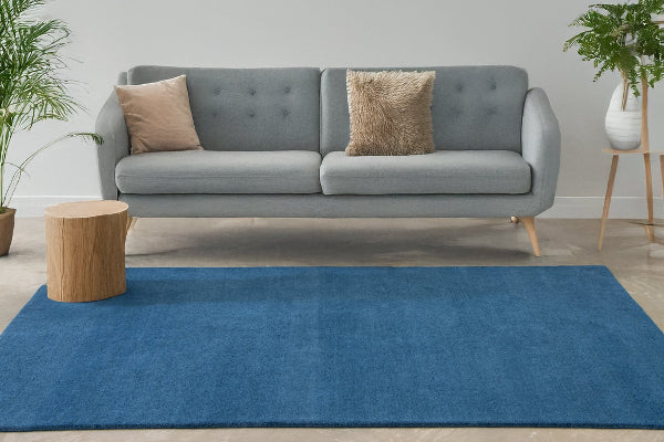 A blue cut pile, hand tufted wool rug placed on the floor in a room with a gray sofa, wooden stool, and potted plant in the background.